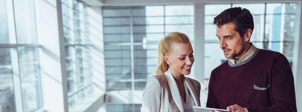 Two business professionals reviewing content on a tablet in a bright, modern office, with colleagues conversing in the background.
