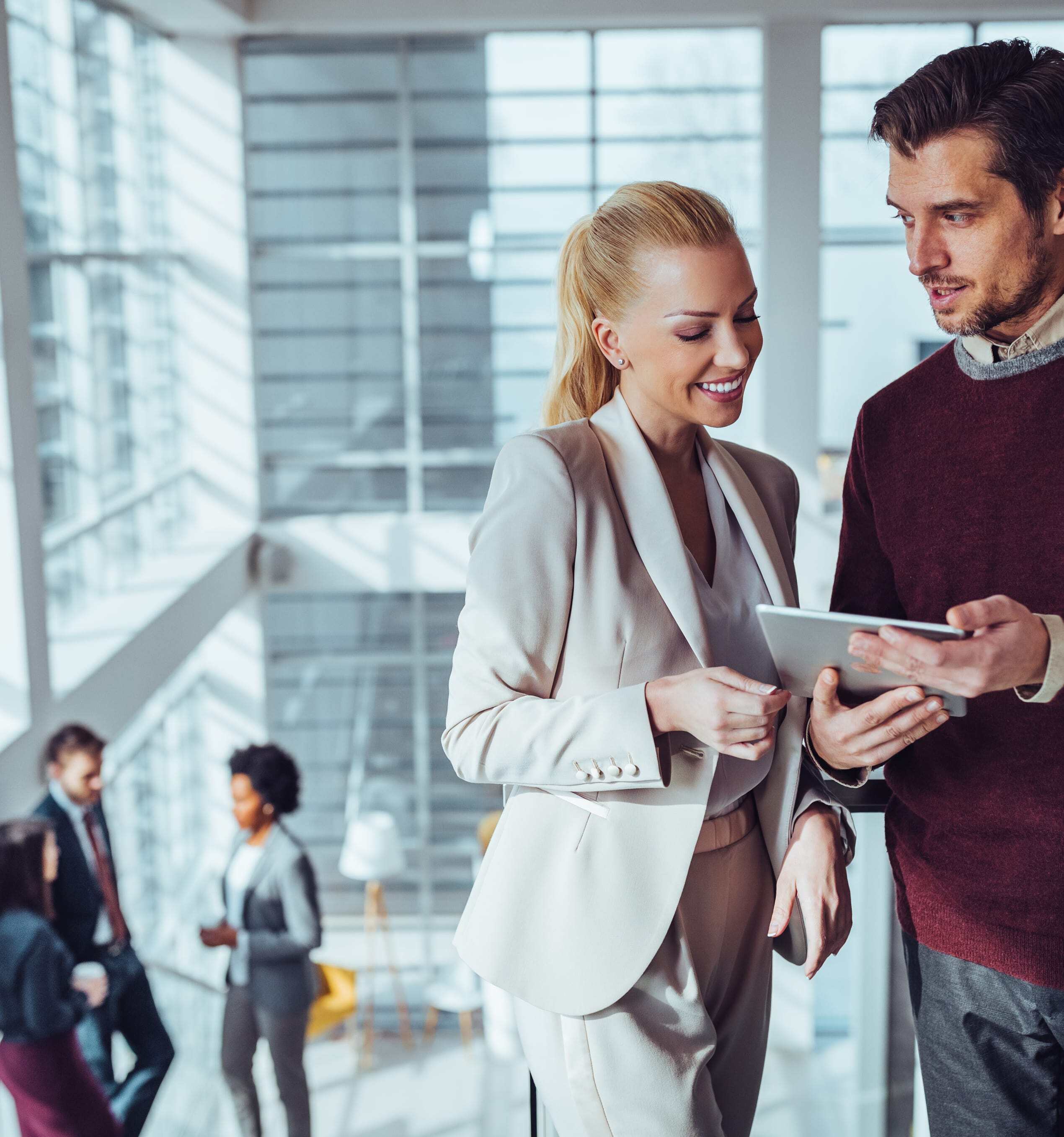 Two business professionals reviewing content on a tablet in a bright, modern office, with colleagues conversing in the background.